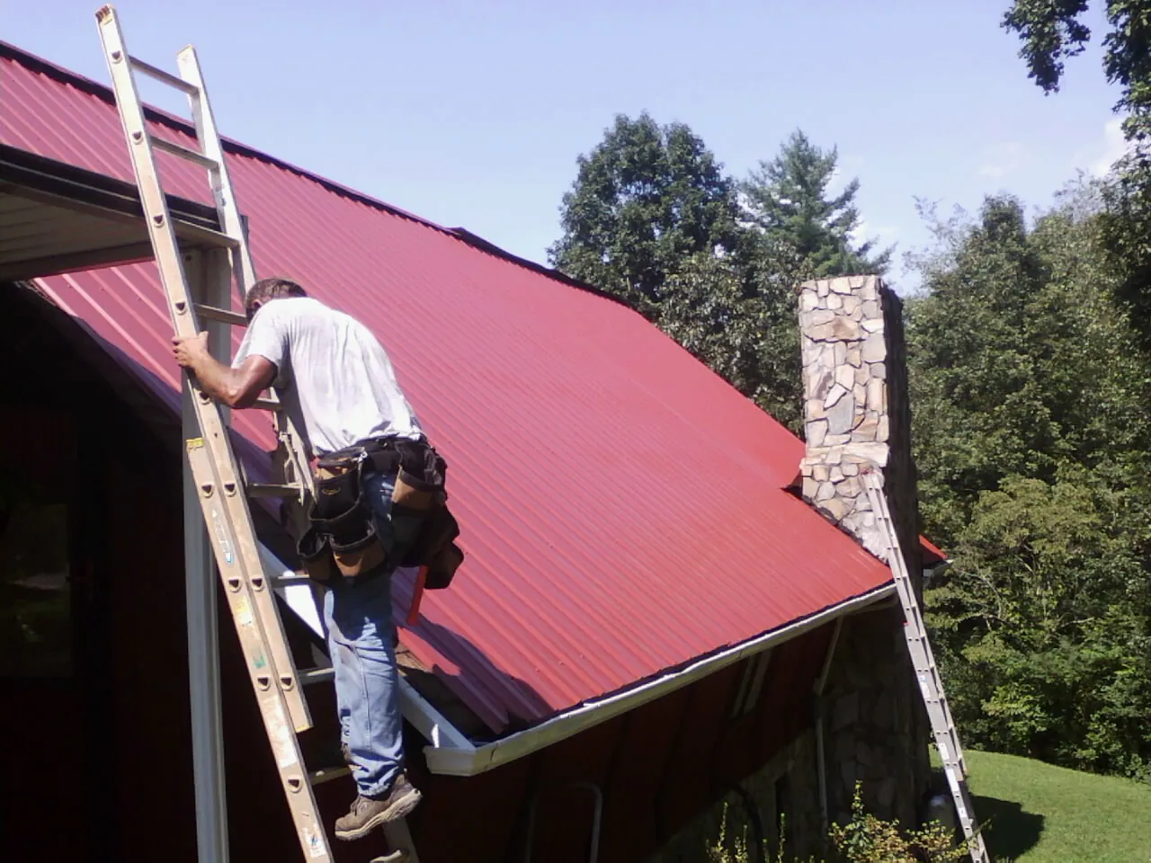 Roofer on a ladder beside a red metal roof and stone chimney — Soffit & Fascia Repair by Roofing Brothers in Amityville